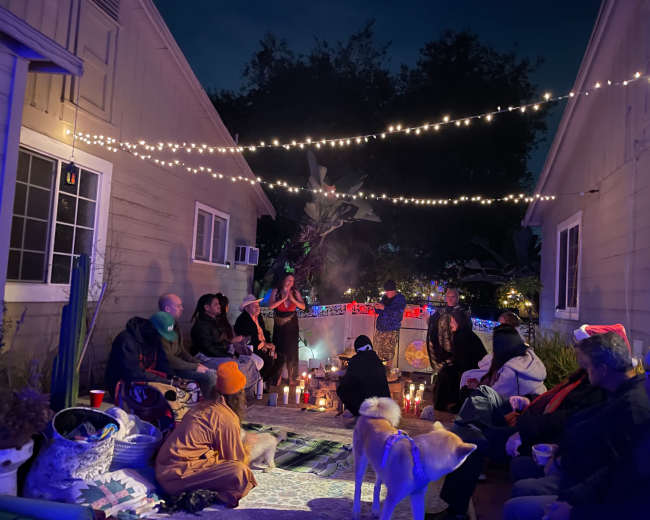 A group of people sits on a patio surrounded by lights and candles, with a performer in the center, during a nighttime gathering.