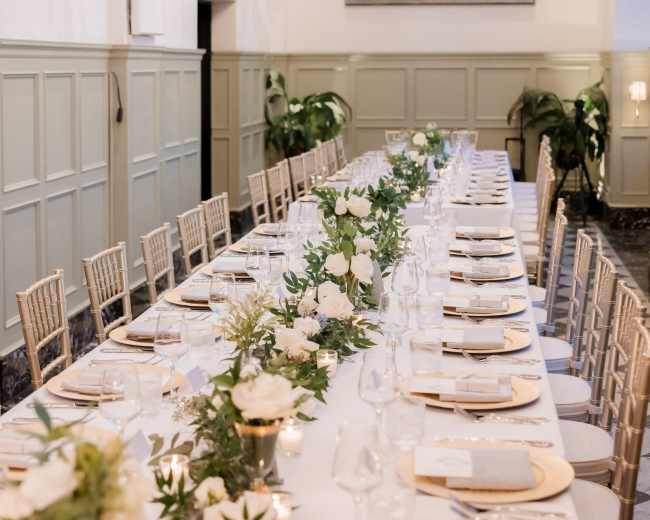 A long banquet table is set with white plates, glassware, and floral arrangements in a elegantly decorated room featuring chandeliers and greenery along the walls.