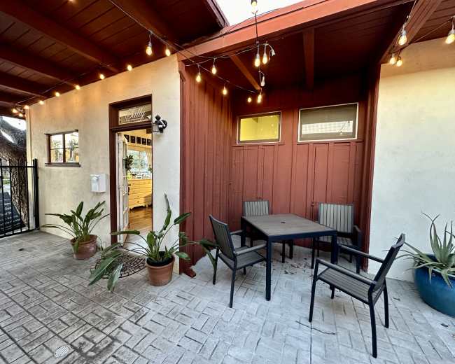 The image shows a small outdoor seating area with a table and chairs, surrounded by potted plants and string lights, adjacent to a building entrance.