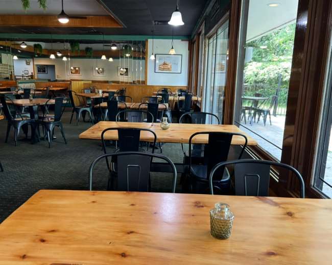 The image shows a spacious, partially empty restaurant with wooden tables and black chairs, featuring large windows that overlook a green outdoor area.