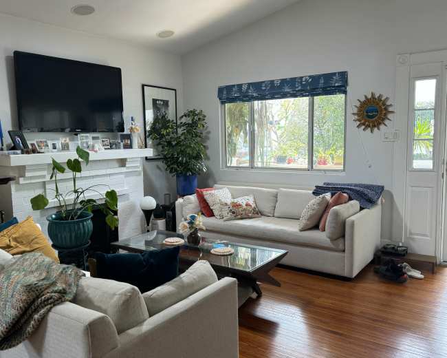 The image shows a living room with two light-colored sofas, a coffee table, a television, and large windows allowing natural light, surrounded by indoor plants and decorative items.