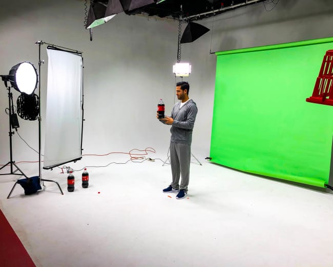 A person stands in a studio with a green screen, holding a bottle of soda, surrounded by lighting equipment and props.