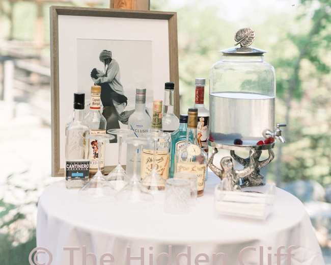 A table draped with a white cloth displays various bottles of liquor alongside a framed black-and-white photograph and a glass beverage dispenser.