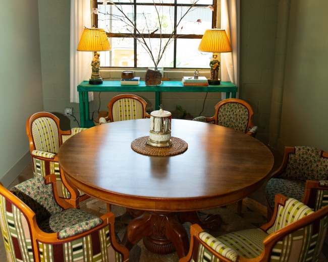 A round wooden dining table with six patterned chairs is set in a well-lit room featuring a large window and decorative lamps.