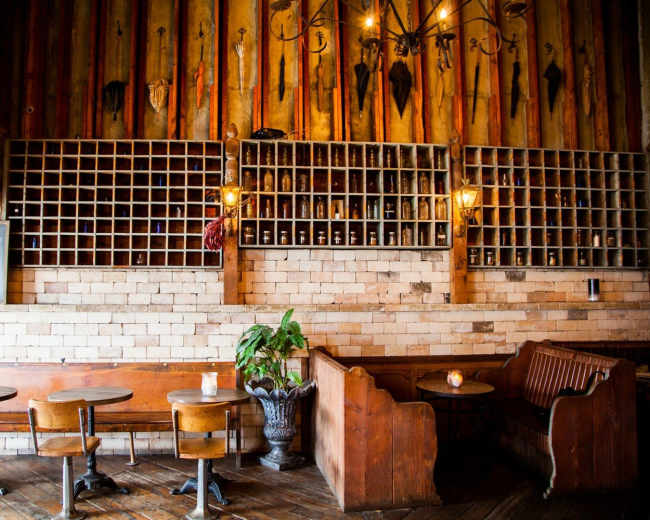 A rustic restaurant features wooden tables and chairs, a wall of shelves displaying bottles, and a decorative arrangement of umbrellas hanging above.