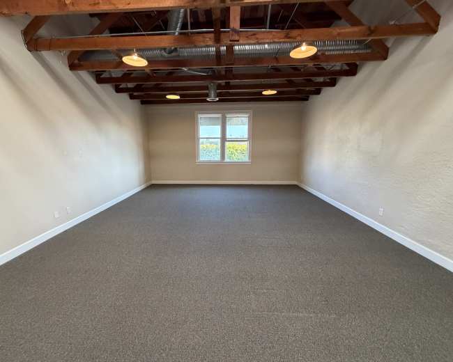 The image shows an empty room with a carpeted floor, exposed wooden beams on the ceiling, and a window allowing natural light to enter.