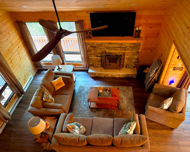 A cozy living room features a large wooden ceiling fan, stone fireplace, and comfortable furniture arranged around a coffee table.
