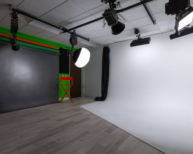 A photography studio with a gray backdrop, a lit area with a white backdrop, two director's chairs, and various lighting equipment.