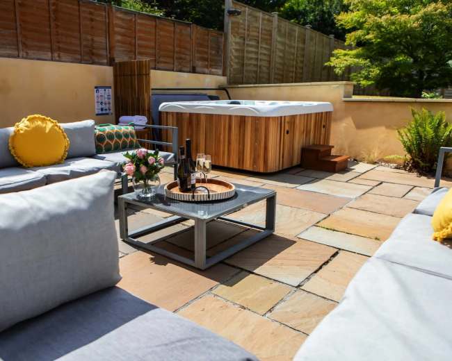 A patio area features a hot tub surrounded by outdoor seating, a coffee table with decorative items, and a backdrop of wood fencing and greenery.