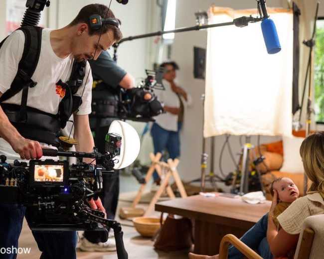 A film crew sets up in a bright room, with a cameraman using a handheld camera to film a mother holding her baby while other crew members adjust equipment.