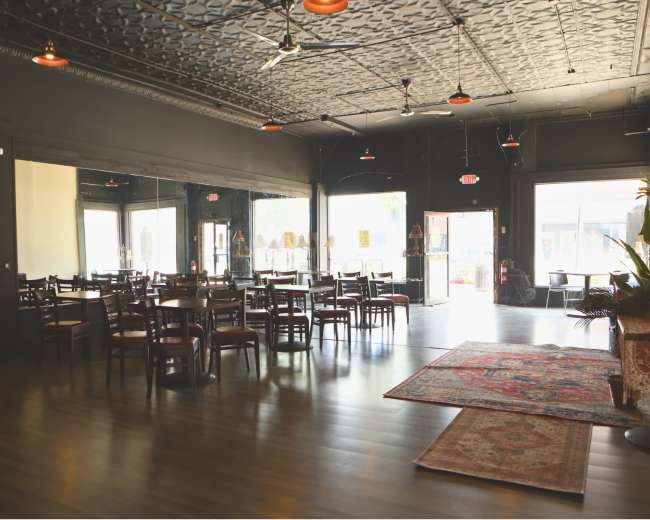 The image shows an empty restaurant interior with wooden tables and chairs, a patterned rug, and large windows letting in natural light.
