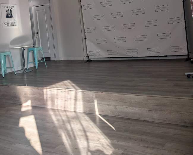 The image shows a bright, minimally decorated room with a plain backdrop on a stand, two teal stools, and wooden flooring.