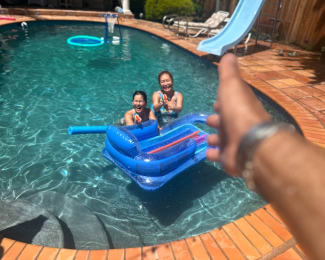 Two children sit on a blue inflatable float in a swimming pool, smiling and holding water guns, while an outstretched hand reaches toward them from the edge of the pool.