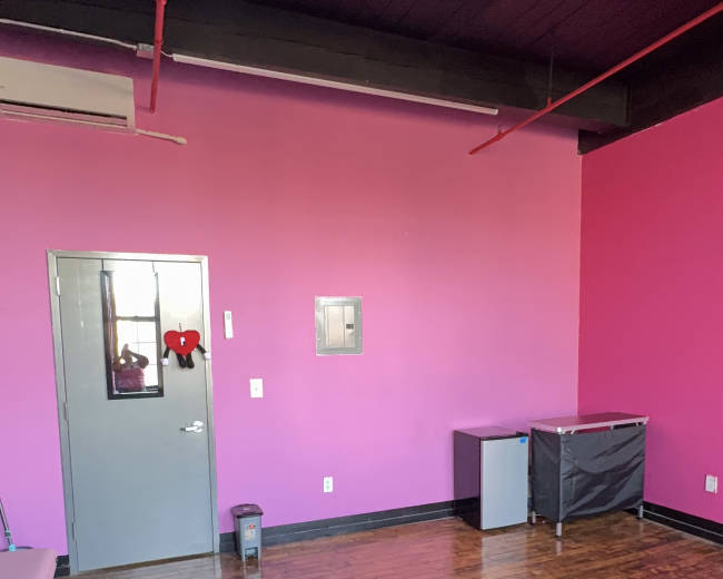 The image shows a room with pink walls, a wooden floor, a door, and a few pieces of furniture including a cabinet and a bin.