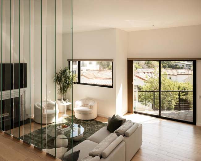 The image shows a modern living room featuring a glass divider, two armchairs, a circular table, and large windows that let in natural light.