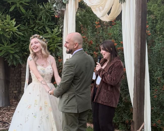 A couple stands in front of a floral-decorated arch while exchanging vows during an outdoor wedding ceremony.