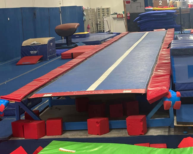 The image shows a gymnastics training area featuring a long trampoline with padded coverings and exercise mats arranged around it.