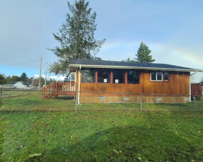 A wooden house with a fenced yard is situated on a grassy lot, with a red car parked nearby.