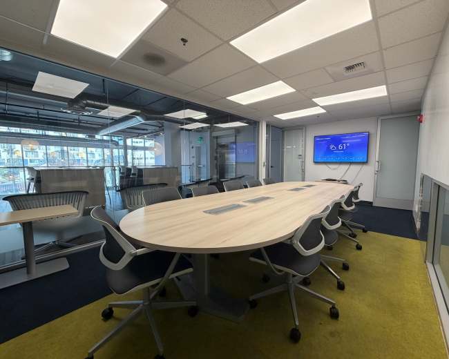 The image shows a modern conference room with a large oval table, ergonomic chairs, and a display screen mounted on the wall.