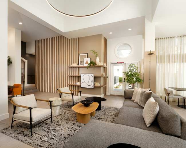 The image shows a modern interior lobby with a gray sectional sofa, wooden accent tables, a minimalist bookshelf, and large windows allowing natural light.