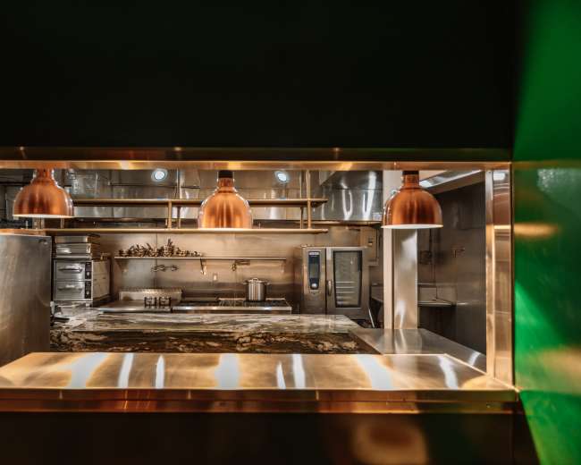 The image shows a stainless steel kitchen visible through a rectangular opening, featuring copper pendant lights and various cooking equipment arranged neatly.