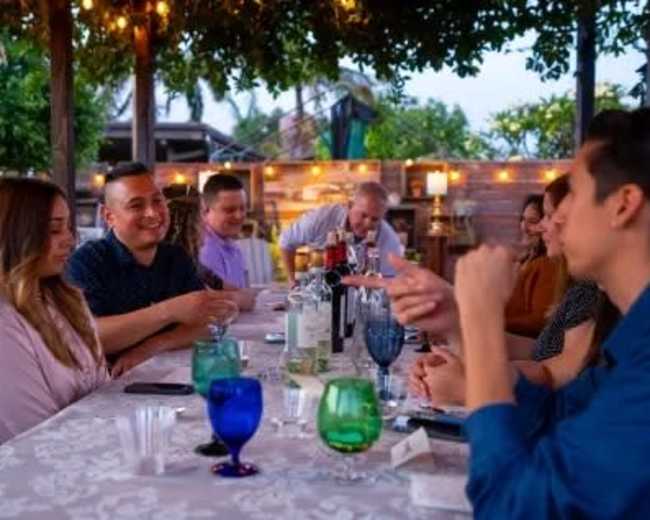 A group of people sits around a table set outdoors, engaged in conversation with drinks and food in the background.