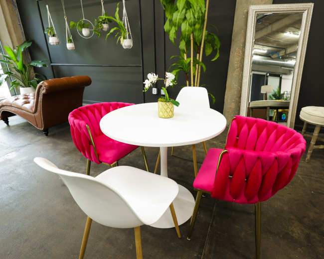 A modern dining area with a round white table surrounded by pink upholstered chairs and a white chair, complemented by plants and a mirror in the background.