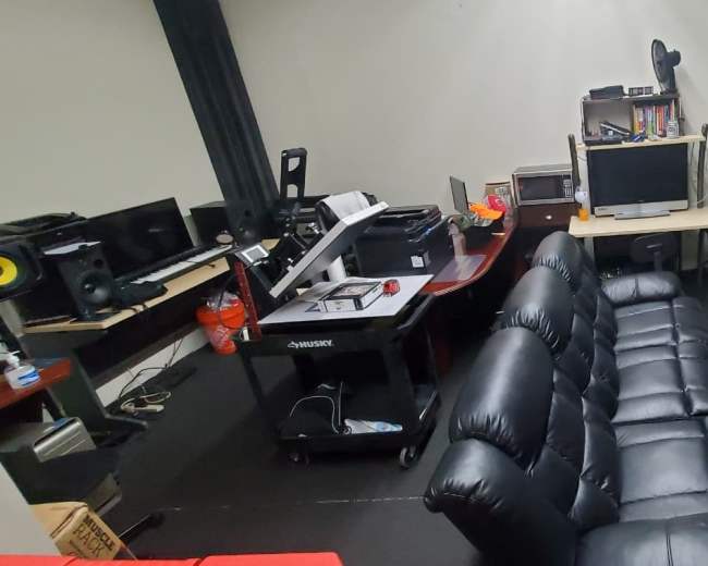 The image shows a recording studio setup with a black leather couch, various audio equipment, a desk with a computer, and lighting equipment positioned in the corner.