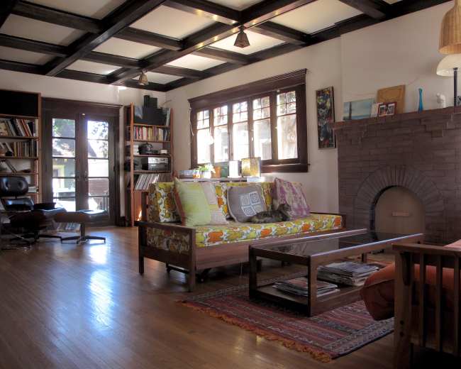 A cozy living room features a patterned sofa, a wooden coffee table with magazines, shelves filled with books, and a brick fireplace under a coffered ceiling.