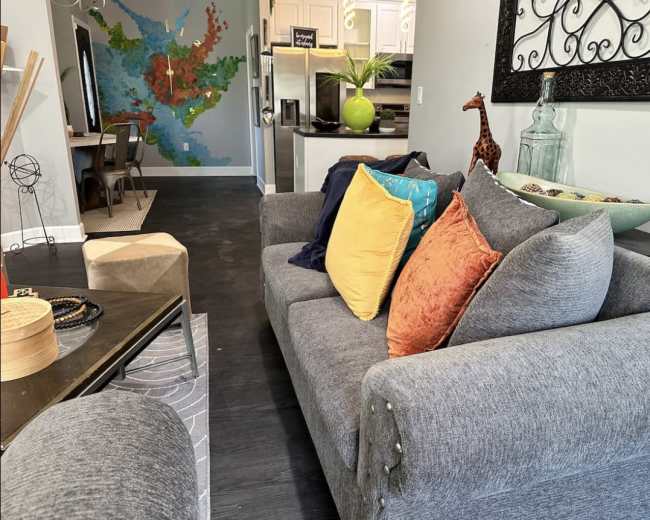 A gray sofa with colorful cushions sits in a living area featuring a map mural on the wall and a dining table in the background.