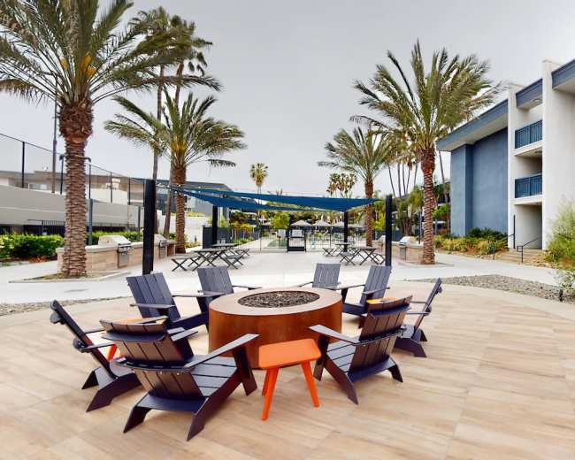 The image shows a landscaped outdoor area with a circular fire pit surrounded by eight wooden chairs, flanked by palm trees and a multi-story building in the background.