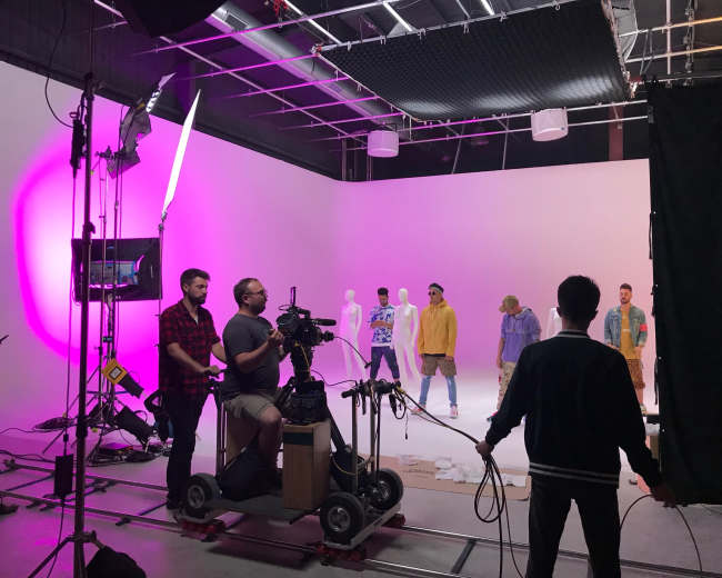 A film crew operates equipment in a studio with models dressed in colorful clothing against a bright pink and white backdrop.