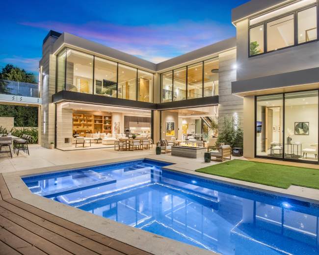 A modern house features large glass windows surrounding a rectangular swimming pool with wooden decking and a neatly landscaped yard.