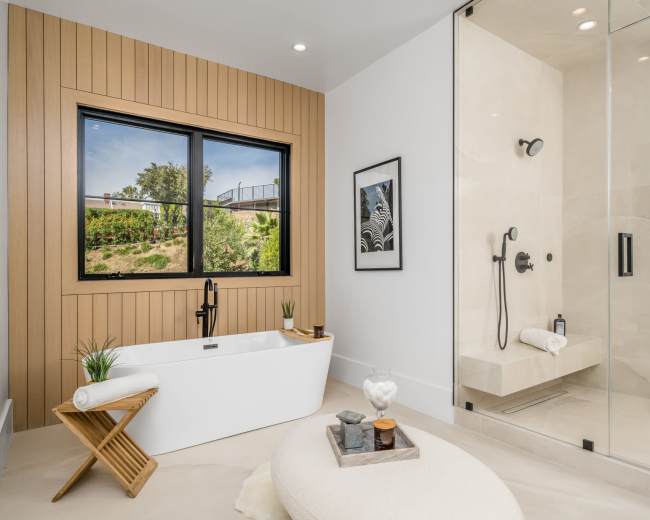 The image shows a modern bathroom featuring a freestanding bathtub, a glass shower enclosure, and large windows with a view of greenery.