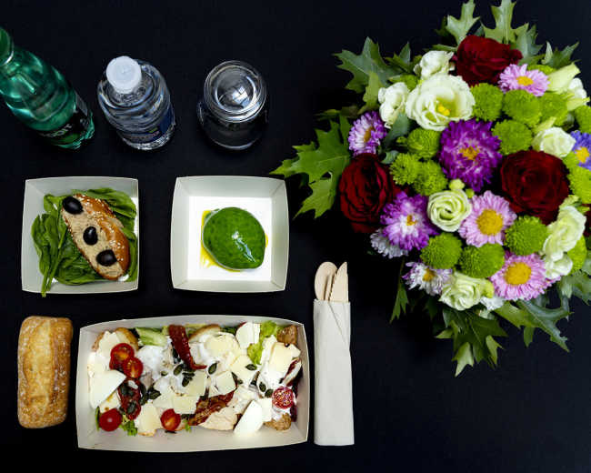 The image features a neatly arranged meal consisting of a box with various salads, a piece of bread with olives, a dessert, bottles of water, and a colorful flower bouquet on a black tablecloth.