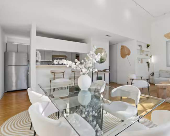 The image shows a bright, modern dining area with a glass table surrounded by white chairs, leading to an open kitchen space.