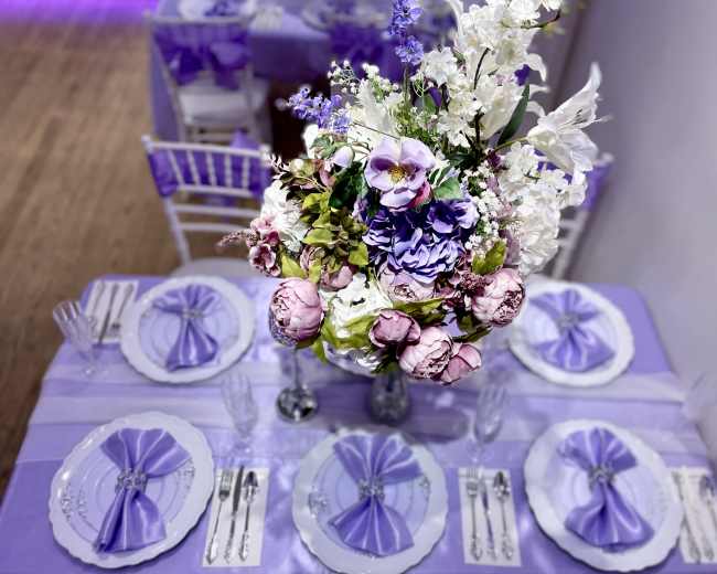 A table set with purple and white decorations features an ornate floral centerpiece and intricately folded napkins.