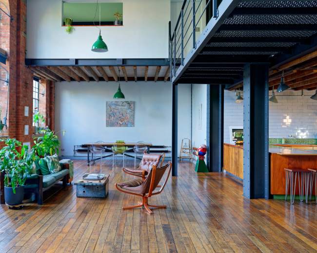 The image shows a spacious, industrial-style loft featuring wooden floors, exposed brick walls, and modern furniture arranged around a central living area.