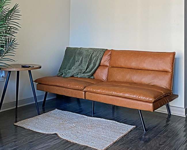A modern living room features a brown leather sofa, a small side table, and two framed leaf prints on the wall.
