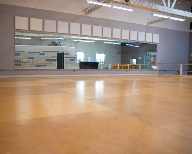 The image shows a spacious dance studio with a polished wooden floor and large mirrors on one wall.