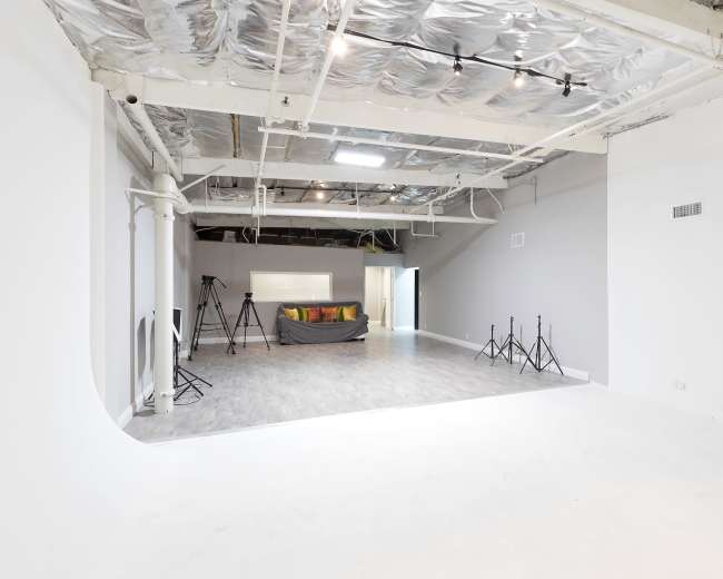 The image shows a spacious, well-lit studio with a gray wall, exposed ceiling, a sofa with decorative pillows, and several tripods set up on the floor.