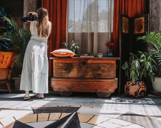 A woman with a camera stands in a room decorated with plants and an antique wooden chest, while natural light streams through a window.