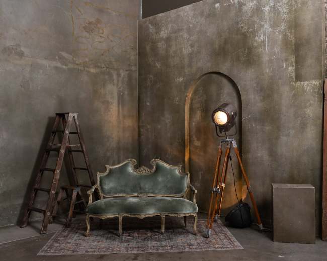 The image shows a vintage green velvet sofa positioned next to a wooden ladder and a large spotlight on a tripod in an unfinished room with textured walls.