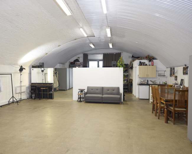 The image shows a spacious room with an arched ceiling featuring a sofa and a dining area with wooden chairs, alongside several cabinets and a kitchenette.