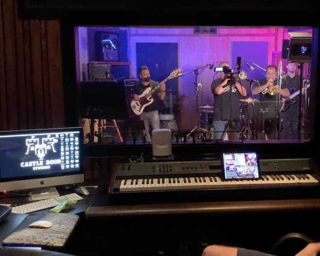 A band performs inside a recording studio while an operator monitors the session from the control room.