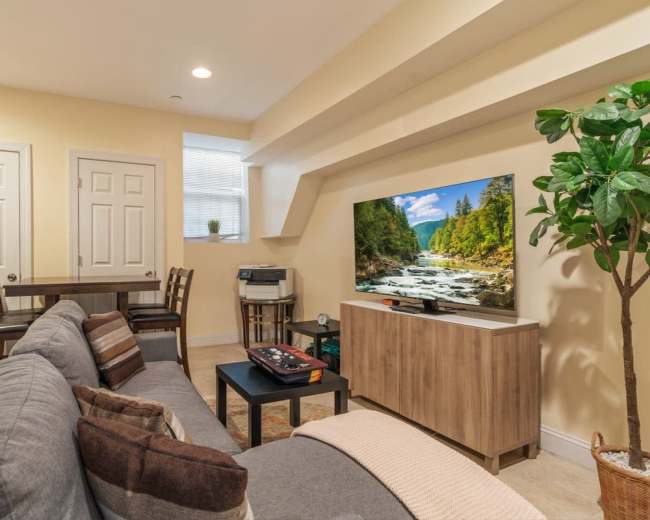 The living room features a sectional sofa, a television mounted on a wooden stand, and a small dining table with two chairs.