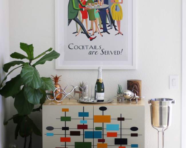 A colorful mid-century modern cabinet stands against a wall featuring a vintage cocktail-themed poster, accompanied by a champagne bottle and a silver serving cart.