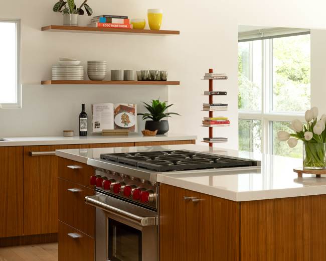 A modern kitchen features wooden cabinetry, a large stainless steel stove with red knobs, and open shelving with decorative items and dishware.