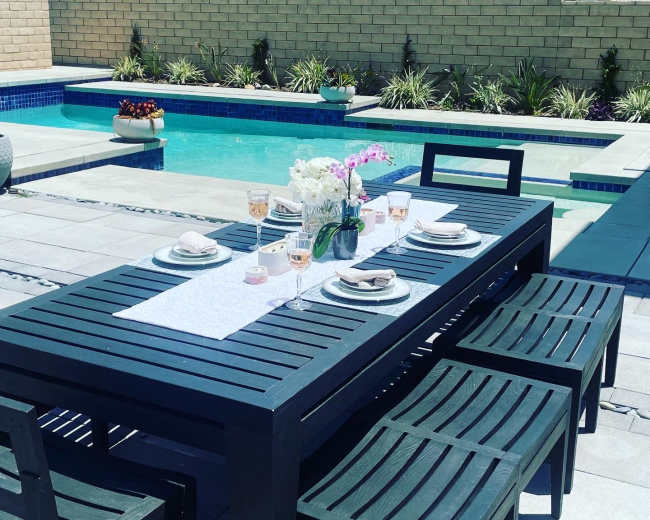 A dining table with place settings and a vase of flowers is set up next to a swimming pool.