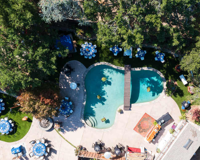 The image shows an aerial view of a landscaped backyard featuring a curvy swimming pool, a wooden bridge, and several tables arranged around it.